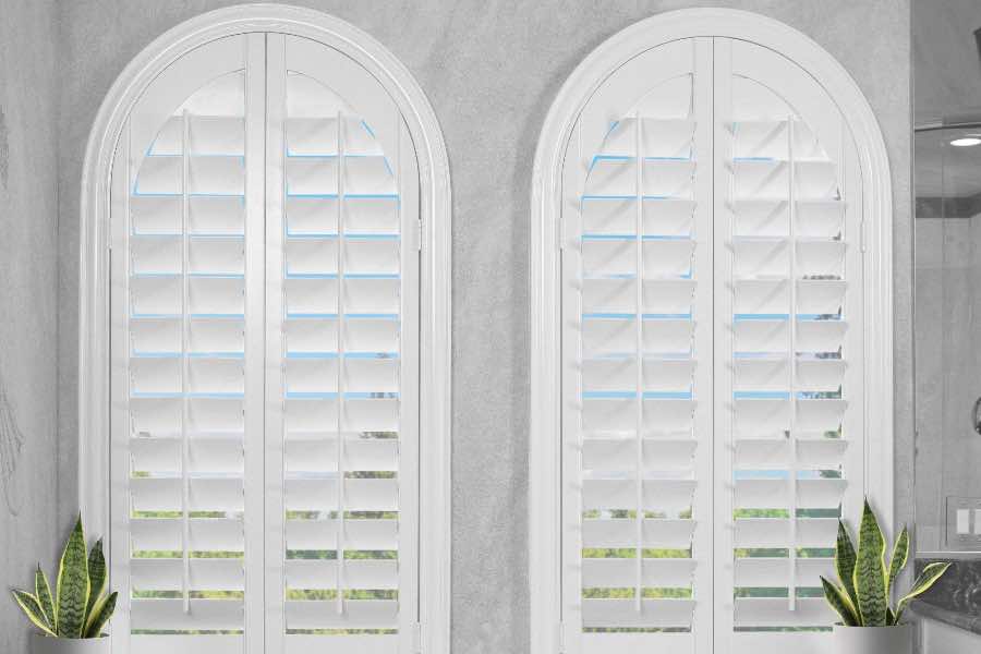 Plantation shutters block light and drafts exceptionally well thanks to their perfect fit. White Polywood shutters on two arched windows by a bathtub