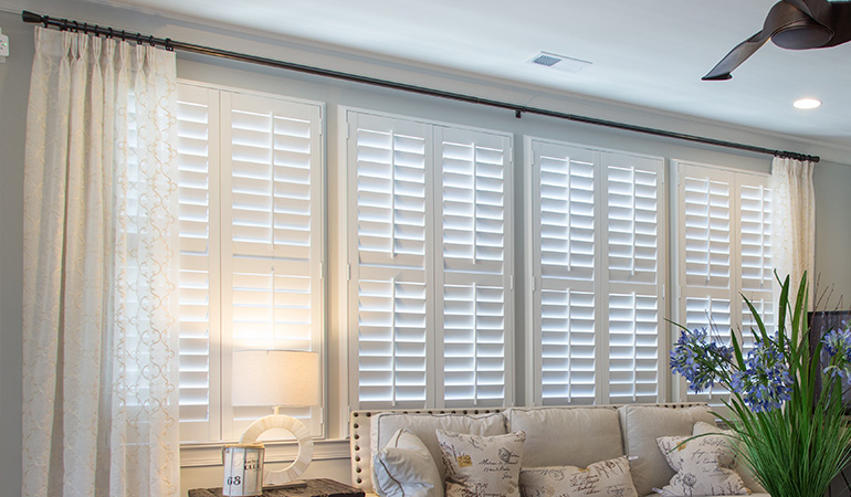 Insulating Polywood shutters paired with energy-efficient windows to reduce energy costs. Large Polywood shutter windows above a white couch inside a living room.